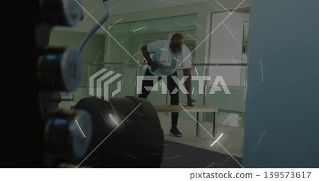 Performing one-arm dumbbell row man wearing light T-shirt, dark pants on bench in gym with towel Performing one-arm dumbbell row man wearing light T-shirt, dark pants on bench in gym with towel 139573617