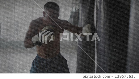 Striking shirtless boxer punching heavy bag at gym, wearing dark shorts, white wraps and gloves Striking shirtless boxer punching heavy bag at gym, wearing dark shorts, white wraps and gloves 139573633