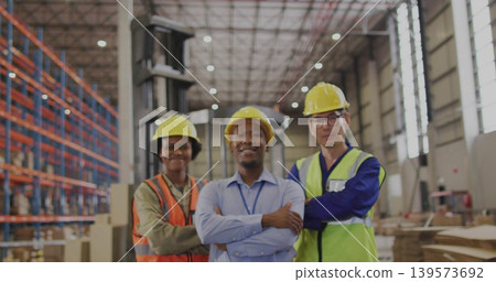 Standing three warehouse workers crossing arms in distribution center, wearing hard hats, vests Standing three warehouse workers crossing arms in distribution center, wearing hard hats, vests 139573692