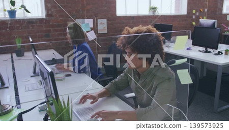 Typing curly-haired man in olive shirt working at open office, with plexiglass dividers and plants Typing curly-haired man in olive shirt working at open office, with plexiglass dividers and plants 139573925