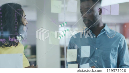 Gesturing coworkers in yellow-top and blue-shirt discussing plan in office, glass with sticky notes 139573928