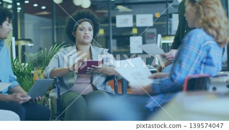 Leading woman in wheelchair in blazer holding tablet, speaking in office with laptop, printouts Leading woman in wheelchair in blazer holding tablet, speaking in office with laptop, printouts 139574047