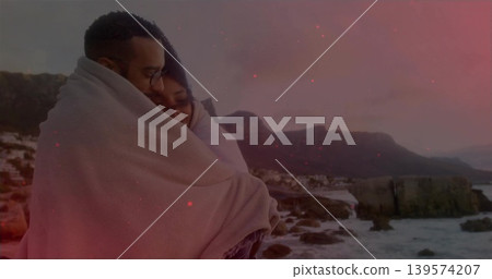 Embracing couple sharing light blanket on rocky shoreline at dusk, man wearing eyeglasses 139574207
