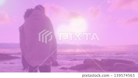 Standing couple wrapped in light blanket facing ocean on rocky shore, pink-purple sun, copy space Standing couple wrapped in light blanket facing ocean on rocky shore, pink-purple sun, copy space 139574563