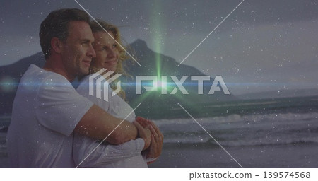 Embracing couple wearing white shirts, gazing at dusk horizon on beach with waves, ridge, flare Embracing couple wearing white shirts, gazing at dusk horizon on beach with waves, ridge, flare 139574568