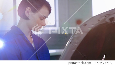 Inspecting woman technician in blue coveralls leaning over car with open hood in garage, lens flare Inspecting woman technician in blue coveralls leaning over car with open hood in garage, lens flare 139574609