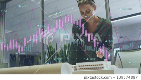 Leaning designer in green sweater holding pen over model at studio with plans, charts, copy space Leaning designer in green sweater holding pen over model at studio with plans, charts, copy space 139574887
