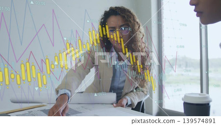 Leaning female professional in blazer pointing at blueprints on conference table, financial overlay Leaning female professional in blazer pointing at blueprints on conference table, financial overlay 139574891