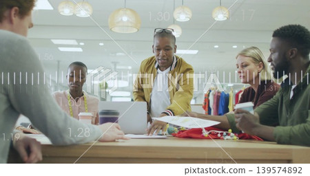 Leaning male team lead wearing mustard jacket pointing at laptop in studio, with fabric swatches 139574892