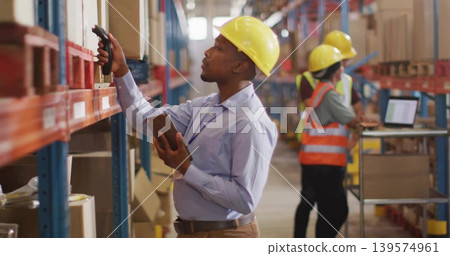 Reaching up warehouse picker wearing yellow helmet scanning box holding tablet and scanner in aisle Reaching up warehouse picker wearing yellow helmet scanning box holding tablet and scanner in aisle 139574961