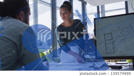 Pointing woman in dark top leaning over table in office, showing plans and screen with overlays Pointing woman in dark top leaning over table in office, showing plans and screen with overlays 139574999