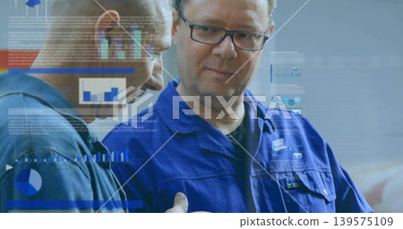 Studying man wearing eyeglasses in blue coverall watching colleague pointing at charts in workshop Studying man wearing eyeglasses in blue coverall watching colleague pointing at charts in workshop 139575109