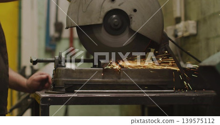 Cutting bench-mounted saw slicing metal workpiece on bench with vise, worker in yellow vest, sparks Cutting bench-mounted saw slicing metal workpiece on bench with vise, worker in yellow vest, sparks 139575112