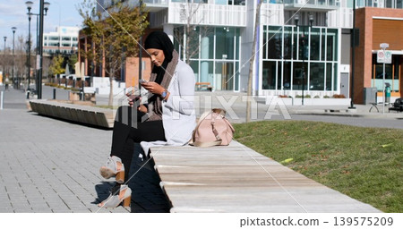 Sitting woman hijab cardigan checking phone holding takeaway cup on bench, pink bag, copy space Sitting woman hijab cardigan checking phone holding takeaway cup on bench, pink bag, copy space 139575209
