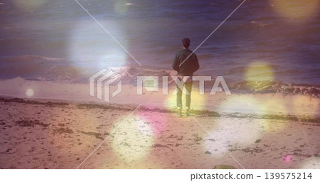 Standing adult man facing ocean on sandy beach, wearing dark jacket, slim jeans, with seaweed line 139575214