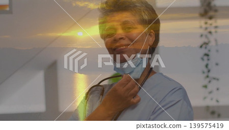 Smiling nurse wearing blue scrubs by window, showing stethoscope and lowered mask with sunset glow Smiling nurse wearing blue scrubs by window, showing stethoscope and lowered mask with sunset glow 139575419
