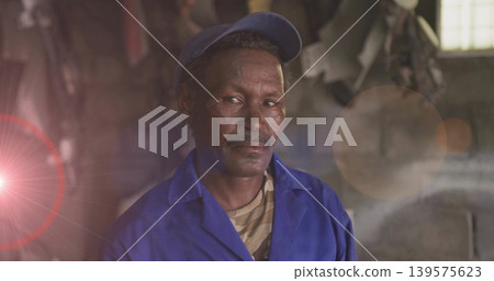 Standing man wearing blue work cap and coveralls in dim workshop, with window grid lens flare Standing man wearing blue work cap and coveralls in dim workshop, with window grid lens flare 139575623
