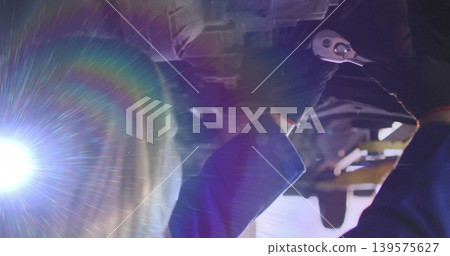 Working tech in coveralls reaching under vehicle on lift in garage, holding ratchet with LED flare Working tech in coveralls reaching under vehicle on lift in garage, holding ratchet with LED flare 139575627