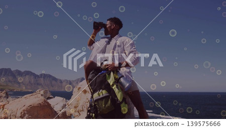 Scanning male hiker wearing light shirt and shorts, on coast, using binoculars and backpack thermos 139575666