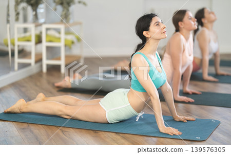 Women bending in back while lying on stomach in Bhujangasana Cobra Stretch position during training together in studio Women bending in back while lying on stomach in Bhujangasana Cobra Stretch position during training together in studio 139576305
