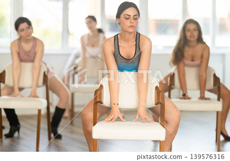Leaning on chair seat with hand foot, young Armenian woman performs lap dance during group workout. 139576316
