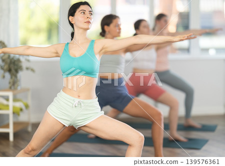 Sporty energetic group of women doing exercises on pilates mat and demonstrating variation of warrior pose of yoga 139576361