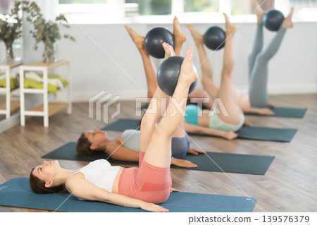 Girl doing leg lifts with ball between feet in group Pilates class Girl doing leg lifts with ball between feet in group Pilates class 139576379