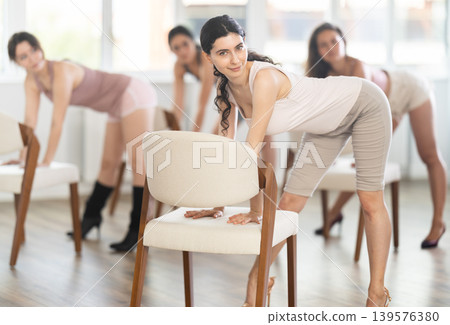 Leaning on chair seat with hand foot, young woman performs lap dance during group workout. 139576380