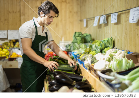 Male salesperson places ripe eggplant on display case. Closeup of salesperson hands and the eggplant. Names of vegetables in Spanish Male salesperson places ripe eggplant on display case. Closeup of salesperson hands and the eggplant. Names of vegetables in Spanish 139576388