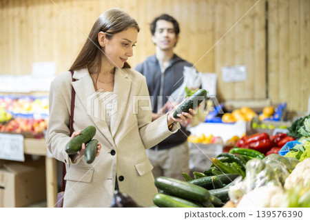 Positive woman chooses fresh cucumbers for purchase in greengrocer shop 139576390
