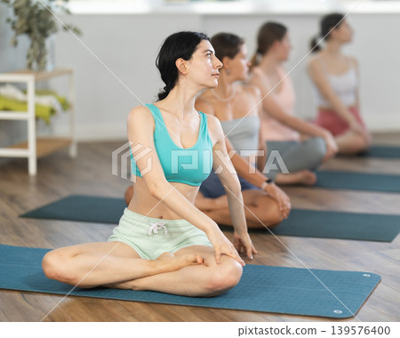 Women maintaining mental and physical health attending group yoga class at modern studio, practicing twisting pose Women maintaining mental and physical health attending group yoga class at modern studio, practicing twisting pose 139576400