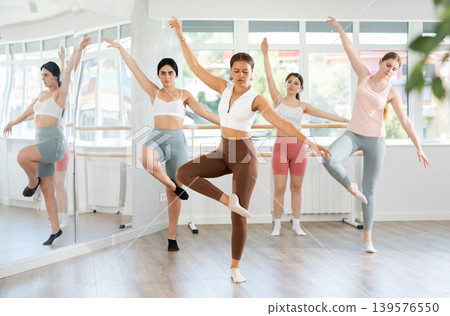 Experienced young girl ballet dancer, dressed in sportswear executing dance moves with focused precision among group of people rehearsing in ballet studio Experienced young girl ballet dancer, dressed in sportswear executing dance moves with focused precision among group of people rehearsing in ballet studio 139576550