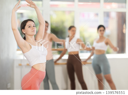Portrait of cheerful active girl exercising ballet moves in fitness studio. Group of young ballet dancers in background near ballet barre 139576551