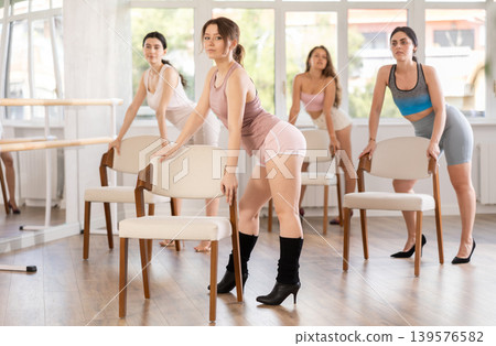 Leaning on chair seat with hand foot, young woman performs lap dance during group workout. 139576582