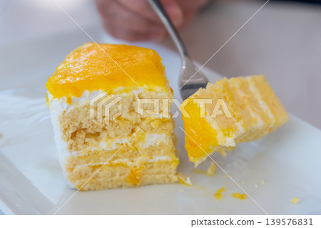 Soft mango cream cake slice with fluffy layers and fresh fruit topping served on a white plate with fork for sweet dessert enjoyment Soft mango cream cake slice with fluffy layers and fresh fruit topping served on a white plate with fork for sweet dessert enjoyment 139576831
