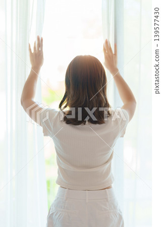 happy woman wake up from a rest and relaxe by the window curtain, young adult female stretching, rising arms and looking to window view in the early morning. healthy lifestyle and morning routine happy woman wake up from a rest and relaxe by the window curtain, young adult female stretching, rising arms and looking to window view in the early morning. healthy lifestyle and morning routine 139577430