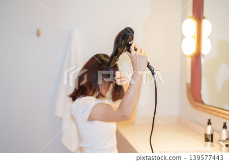 Young woman drying long hair with a hair dryer while looking at the mirror after a shower in the bathroom at home or hotel room during vacation. Hairstyles and daily morning routine Young woman drying long hair with a hair dryer while looking at the mirror after a shower in the bathroom at home or hotel room during vacation. Hairstyles and daily morning routine 139577443