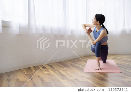 一位中年日本女性在明亮的窗邊，在瑜珈墊上做著瑜珈扭轉式。 139578341