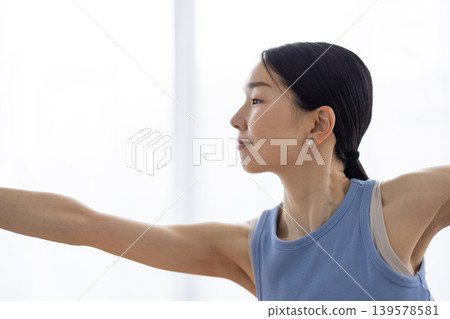 A middle-aged Japanese woman strikes a dancer's pose on a mat by a bright window. A middle-aged Japanese woman strikes a dancer's pose on a mat by a bright window. 139578581
