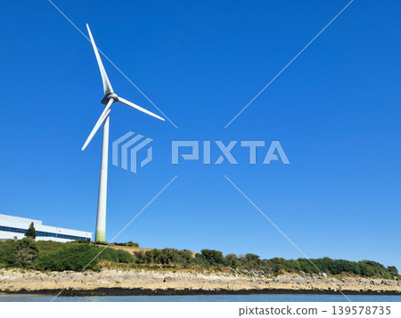 Wind turbine beside rocky shoreline under blue sky 139578735