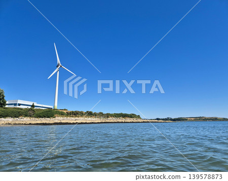 Wind turbine by calm coastal water under blue sky Wind turbine by calm coastal water under blue sky 139578873