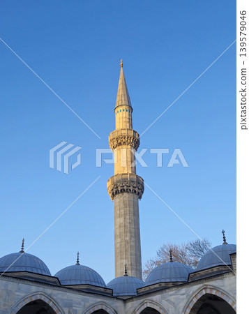 Tall mosque minaret above domed roof against blue sky Tall mosque minaret above domed roof against blue sky 139579046