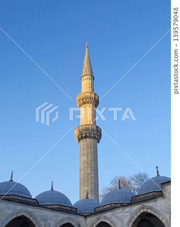 Tall mosque minaret above domed roof against blue sky Tall mosque minaret above domed roof against blue sky 139579048