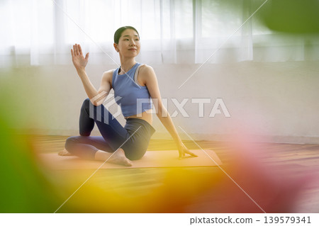 A middle-aged Japanese woman performs the Pisces pose in yoga on a mat by a bright window. A middle-aged Japanese woman performs the Pisces pose in yoga on a mat by a bright window. 139579341