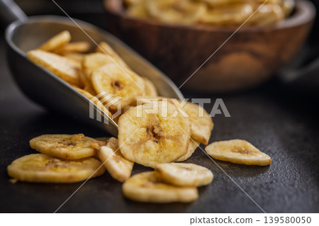 Crispy Banana Chips in Metal Scoop and Wooden Bowl on Dark Surface. Dried Fruit Snack Crispy Banana Chips in Metal Scoop and Wooden Bowl on Dark Surface. Dried Fruit Snack 139580050