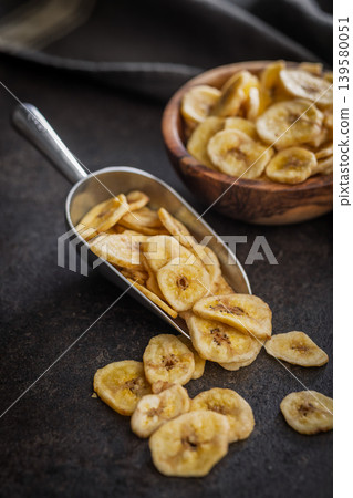 Crispy Banana Chips in Metal Scoop and Wooden Bowl on Dark Surface. Dried Fruit Snack Crispy Banana Chips in Metal Scoop and Wooden Bowl on Dark Surface. Dried Fruit Snack 139580051