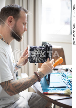 Man works on computer hardware assembly in a well-lit room during daytime in a home setting Man works on computer hardware assembly in a well-lit room during daytime in a home setting 139582882