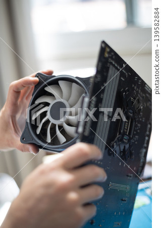 Person installs a computer fan onto a circuit board in a workspace during daytime hours Person installs a computer fan onto a circuit board in a workspace during daytime hours 139582884