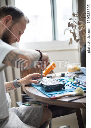 Man works on repairing electronics at home during the day with tools and materials scattered around the table Man works on repairing electronics at home during the day with tools and materials scattered around the table 139582895