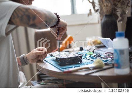 Person repairs a small electronic device on a table at home in daylight while tools and materials are nearby Person repairs a small electronic device on a table at home in daylight while tools and materials are nearby 139582896